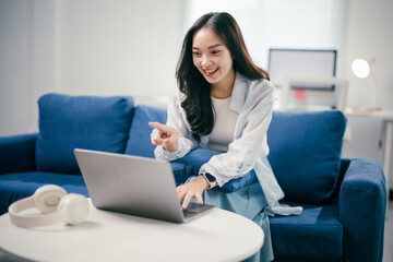 Young asian freelancer woman is sitting on a blue couch and pointing her finger at a laptop screen while working remotely from her home office