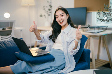 Fototapeta premium Young woman happily works on her laptop in a modern living room, giving two thumbs up