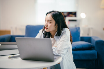 Young asian woman is sitting on the floor in her living room, working at home on her laptop. She is staring thoughtfully into the distance, taking a break from her computer work