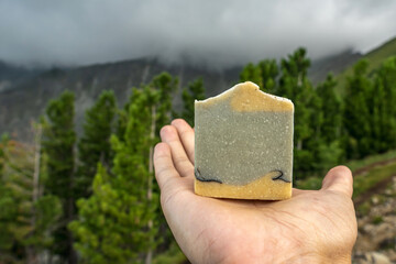 Piece of craft soap in a hand against the background of mountains, forests and sky. Concept of the image of organic cosmetics in a natural environment