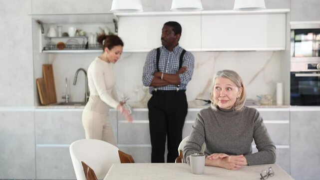 Elderly woman smiling during family quarrel between couple of adult man and woman in kitchen