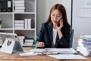 Attractive business asian woman secretary in office working on laptop documents paperwork and talking to someone on mobile phone share good news about project in office workplace,
