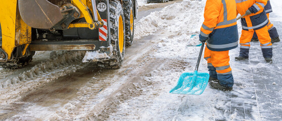 Two men in orange and blue work together to clear snow from a road