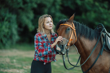 Obraz premium Happy blonde with horse in forest. Woman and a horse walking through the field during the day. Dressed in a plaid shirt and black leggings.