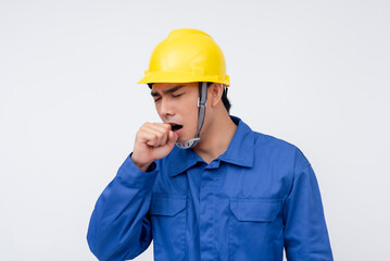 A young construction worker in blue coveralls and a yellow hard hat is yawning, appearing tired. Isolated on a white background, the image conveys fatigue and hard work.