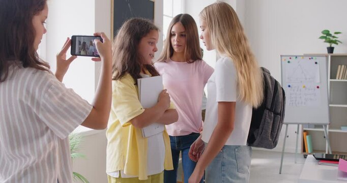 Bullying in educational environment. Group of teenage children bully girl in school and capture it on smartphone camera. One child is holding mobile phone, recording how two others surround girl.