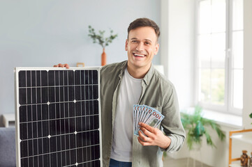 Portrait of a happy, smiling, joyful man at home holding a photovoltaic solar panel and a fan of US dollar bills. Concept of using sustainable technology and clean solar energy to save money © Studio Romantic