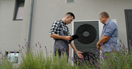 A technician consults with a homeowner about a heat pump, using a tablet for demonstration beside the unit.