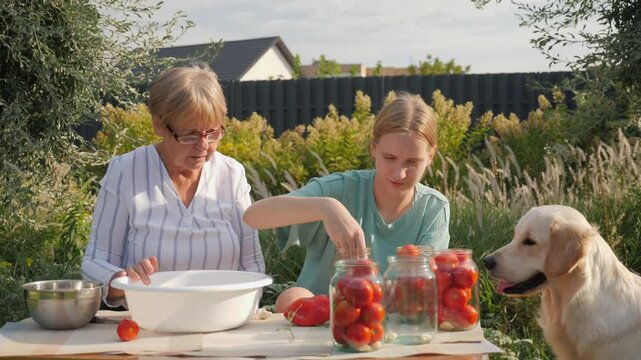 A grandmother and her granddaughter are canning tomatoes in a serene garden, with their dog resting beside them.