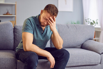 Stressed upset man sitting alone on a comfortable couch, appearing distraught, holding his head in hands, deep in thought or contemplation. Sad guy thinking about problems in relationships or at work