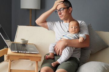 Exhausted father touching his forehead working on his laptop from the couch trying to manage remote business with taking care of his infant baby feeling the strain of parenting and online work