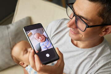 Dad in glasses holding his adorable newborn daughter or son in his hands while taking photo with mobile phone indoors creating beautiful memory in the nursery