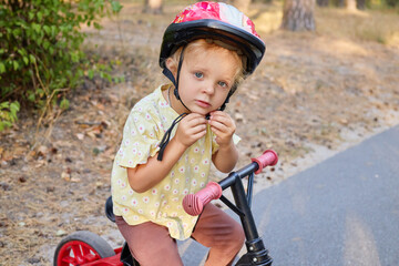 Naklejka premium Little wavy haired girl balancing on her bike along the path in park enjoying the warmth of the sun and the thrill of her first outdoor adventure with push bike