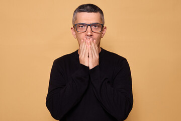 Nervous adult male wearing black jumper and glasses looking at camera with anxiety and frustration has frightened expression feeling scared and confused isolated over beige background