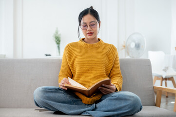 A beautiful Asian woman in casual clothes focuses on reading a book on a couch at home.