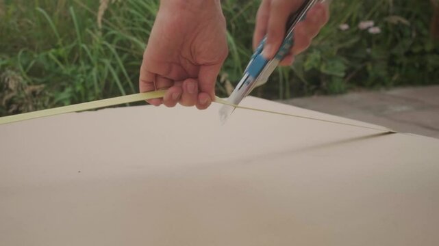 Handheld shot of opening a big package outside on the terrace. Female caucasian hands holding a box cutter and opening the string. Camera follows the action.