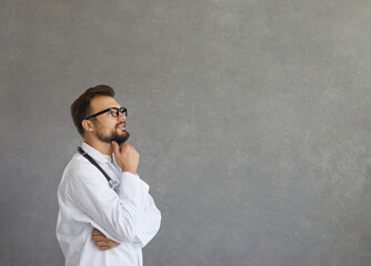 Smiling dreamy pensive young male caucasian doctor in white medical uniform look in distance. Medical worker thinking or pondering, planning future career or success in medicine scratching beard