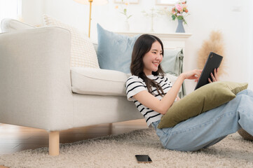 Fototapeta premium A beautiful woman sits on a large, comfortable sofa by the window of her house. She plays social media on tablet during the weekend.