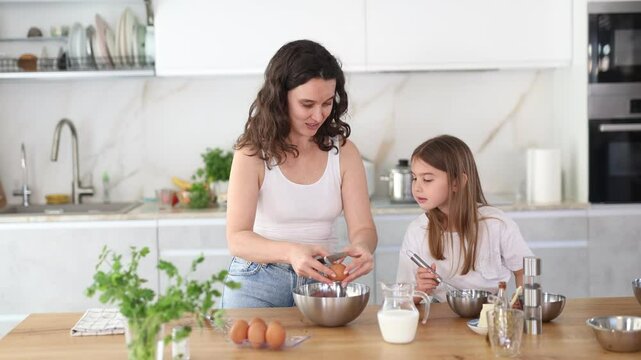 Mom teaches her daughter how to make dough, breaks eggs into bowl. Woman explains to her daughter successful recipe and rules for making dough for dessert. Homemade food and little helper.