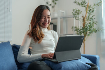 Fototapeta premium Attractive Asian woman sitting on sofa in living room. She uses laptop, tablet to play social media and chat online with friends, work contact, learn work. Concept of using technology.