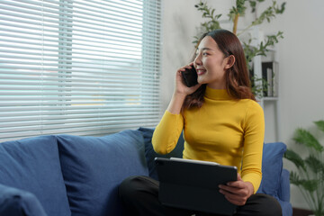 Happy attractive Asian woman holding mobile phone using technology for online shopping, e-commerce, buying things online, texting, greeting, chatting, relaxing on sofa in living room at home.