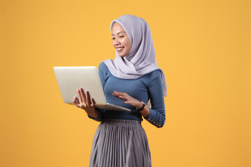 happy asian indonesian muslim woman holding laptop computer on isolated yellow background