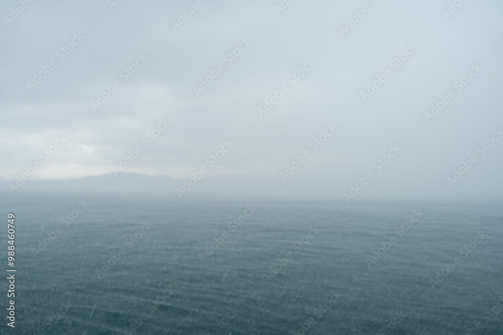 Poster light rain creates ripples on the surface of the sea while an overcast sky looms over a tranquil coa