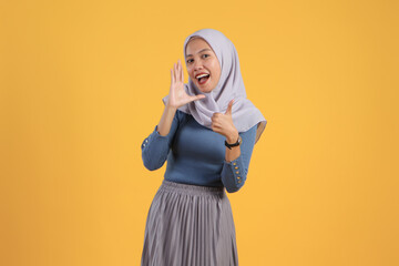 asian indonesian muslim woman shouting and giving thumb up finger gesture on isolated yellow bakcground