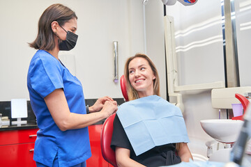 A woman is sitting in a dentist's chair with a blue cloth over her mouth. A dental assistant is...