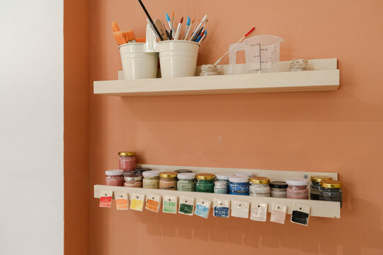 Craft products and water color paints kept in jars on shelf over peach wall