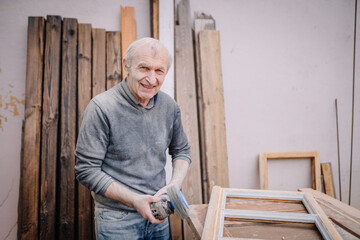 An elderly man smiling while sanding a wooden frame in a workshop filled with wooden planks and tools..