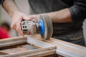 Close-up of a man’s hands using a power sander to smooth a wooden window frame. The image captures the details of the tool and manual work..