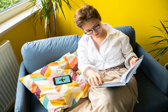 Woman reading book with flight mode symbol on smart phone at home