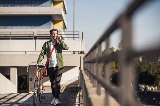Happy man talking on smart phone and walking with bicycle in front of building