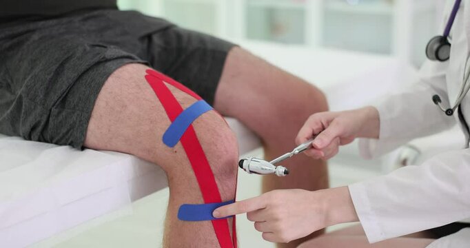 Doctor tests nerve response using reflex hammer checking knee. Patient with kinesiology stretch tape undergoes examination in private clinic