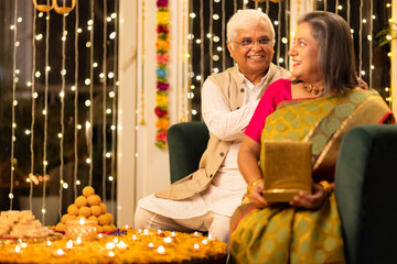 Happy senior husband giving surprise gift to excited wife in illuminated home on occasion of Diwali festival