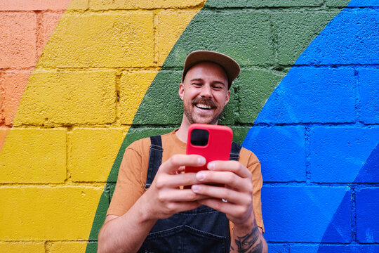 Happy gay man using smart phone and standing in front of colored wall