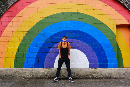 Playful gay man with hands in pockets standing in front of rainbow wall