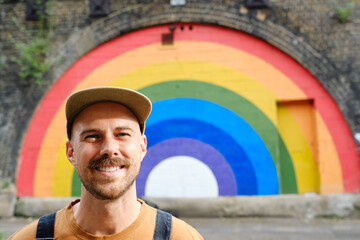 Smiling gay person wearing cap near rainbow painted wall