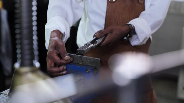 A male jeweller drawing wire through a metal block with pliers to make the wire taut and surrounded by machinery