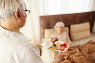 Rear view of senior man bringing breakfast in bed to his wife.