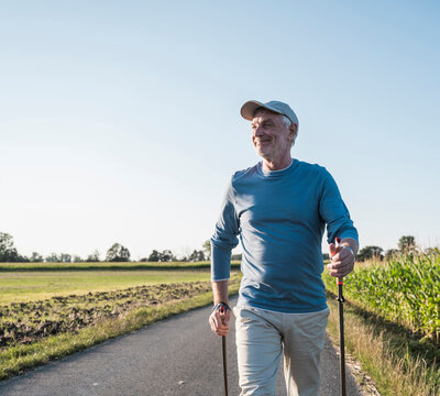 Smiling senior man holding hiking poles and walking on road near field