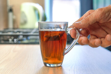 Someones hand is holding a glass mug of tea with a teabag in a kitchen environment