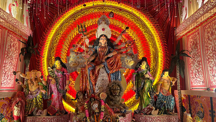 A powerful depiction of Maa Durga, the fierce Hindu goddess, with multiple arms holding divine weapons. She stands confidently on a lion, symbolizing strength and protection, dressed in vibrant red an