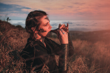Beautiful girl in gothic style clothes with tattoo on her face plays ocarina in the mountains