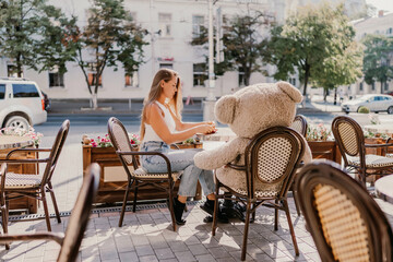 A woman sits cafe with a teddy bear next to her. The scene is set in a city with several chairs and tables around her. The woman is enjoying her time at the outdoor cafe.