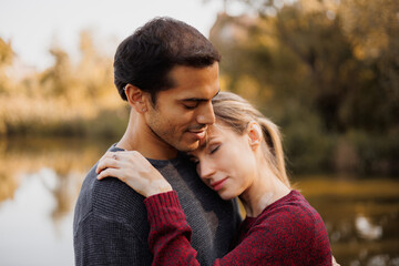 Portrait of woman in sweater hugging mixed race boyfriend near blurred lake