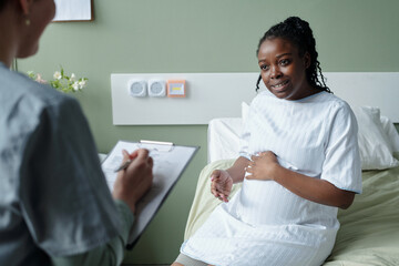 Expectant mother sitting on hospital bed wearing hospital gown and smiling warmly discussing with...