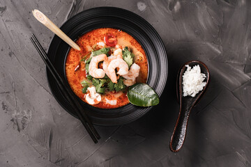 Tom Yam Kung Spicy Thai soup with shrimp, lemongrass and galangal in a black bowl on a dark background and a spoon with rice. top view