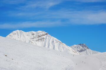 Snowy slopes with ski riders against mountain range in sunny day. Ski resort Gudauri, Georgia. Caucasus Mountains. Aerial view.
